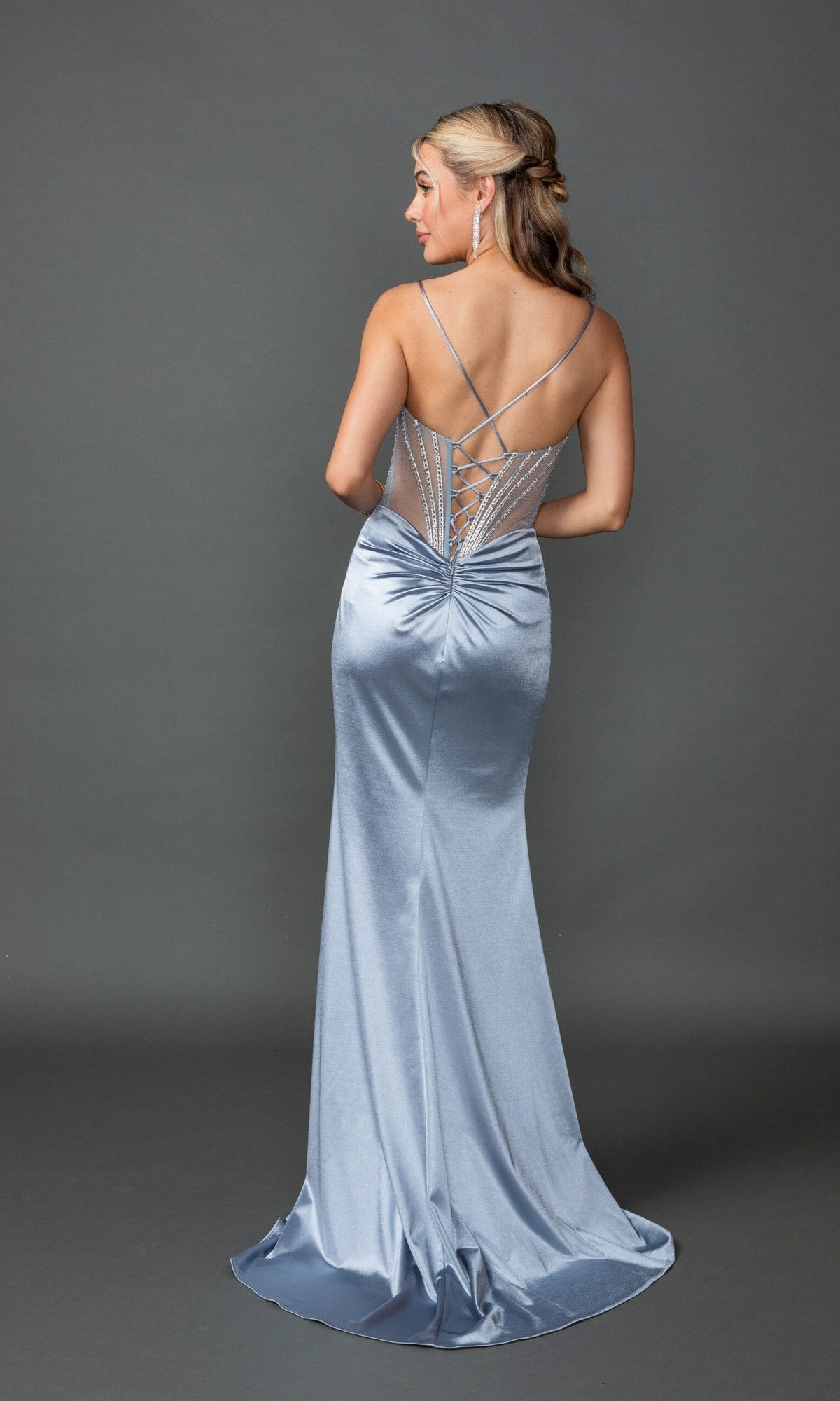 Woman in the Dancing Queen 4538 long prom dress, silver-blue satin with lace-up back and thin straps, stands facing away against a gray background.